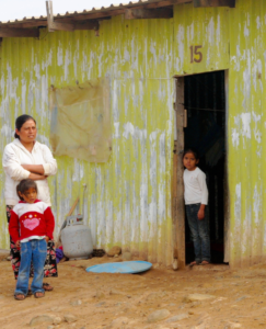 A woman stands with crossed arms beside a child in a red sweater, while another child peers from the doorway of a weathered green house with the number 15