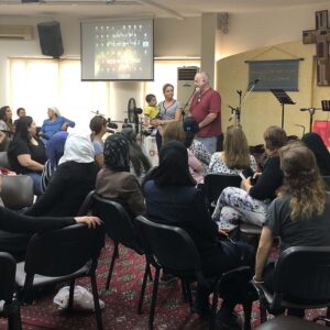 A diverse group of people seated inside a room with a man, woman, and child standing in front, as part of a ministry gathering in Lebanon