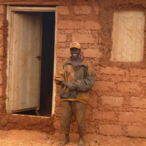 A smiling man standing beside a mud-brick house with a metal door in Burundi