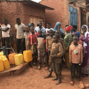 Busy scene in Burundi showing people of various ages standing near yellow water containers outside a red brick building