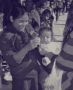 A blurred image of a woman holding a baby and a small container, with other people in the background, likely at an outdoor gathering in Mexico