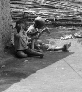 Two children sitting by a mud wall, playing with simple toys, in a rural village setting