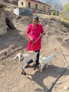 A smiling Nepali woman in a bright pink dress holding a goat on a leash with stone houses in the background.