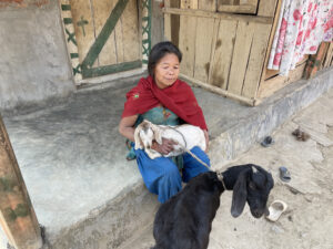 A Nepali woman in a red shawl and blue dress seated with goats in a village home setting.